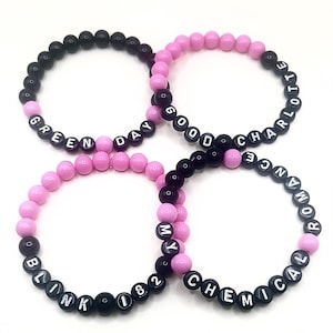 May include: Four circular beaded bracelets featuring black and pink spherical beads. The bracelets display white letter beads spelling out the band names Green Day, Good Charlotte, Blink 182, and My Chemical Romance in a repeating pattern.