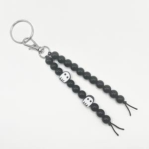 May include: A keyring featuring a silver metal ring and lobster clasp attached to two strands of black beads. Each strand includes a white ghost-shaped bead with black eyes and a mouth. The strands are finished with short black cord ties at the ends.