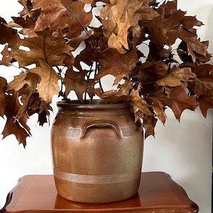 Peut inclure: Un pot en grès brun avec deux poignées contient un bouquet de feuilles de chêne brunes séchées. Le pot est posé sur une petite table en bois foncé. Les feuilles sont marron clair et foncé.