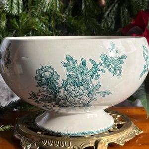 Puede incluir: Un cuenco de cerámica blanca con diseños florales turquesas y asas. El cuenco se encuentra sobre un soporte decorativo de latón. El patrón floral incluye grandes flores parecidas a peonías y follaje.