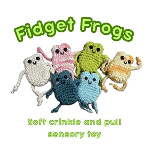 May include: A collection of six crocheted frog toys in various colors, including white, pink, green, blue, and yellow. The toys have black eyes and are arranged on a white background. Text reads "Fidget Frogs" and "Soft crinkle and pull sensory toy."