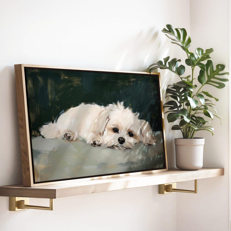 May include: A framed canvas art print featuring a white dog lying down. The dog has a fluffy coat and dark eyes. The background is a dark green color. The frame is made of light-colored wood. The artwork is displayed on a wooden shelf.