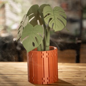 May include: A decorative plant with large, green leaves in a terracotta-colored pot. The pot has a geometric design. The plant is a Monstera, also known as a Swiss cheese plant, and is displayed on a wooden surface.