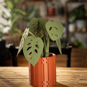 May include: A decorative artificial plant with green leaves and a terracotta-colored pot. The plant features large, perforated leaves and is set in a cylindrical pot with a geometric design. The pot is made of a reddish-brown material and sits on a wooden surface.