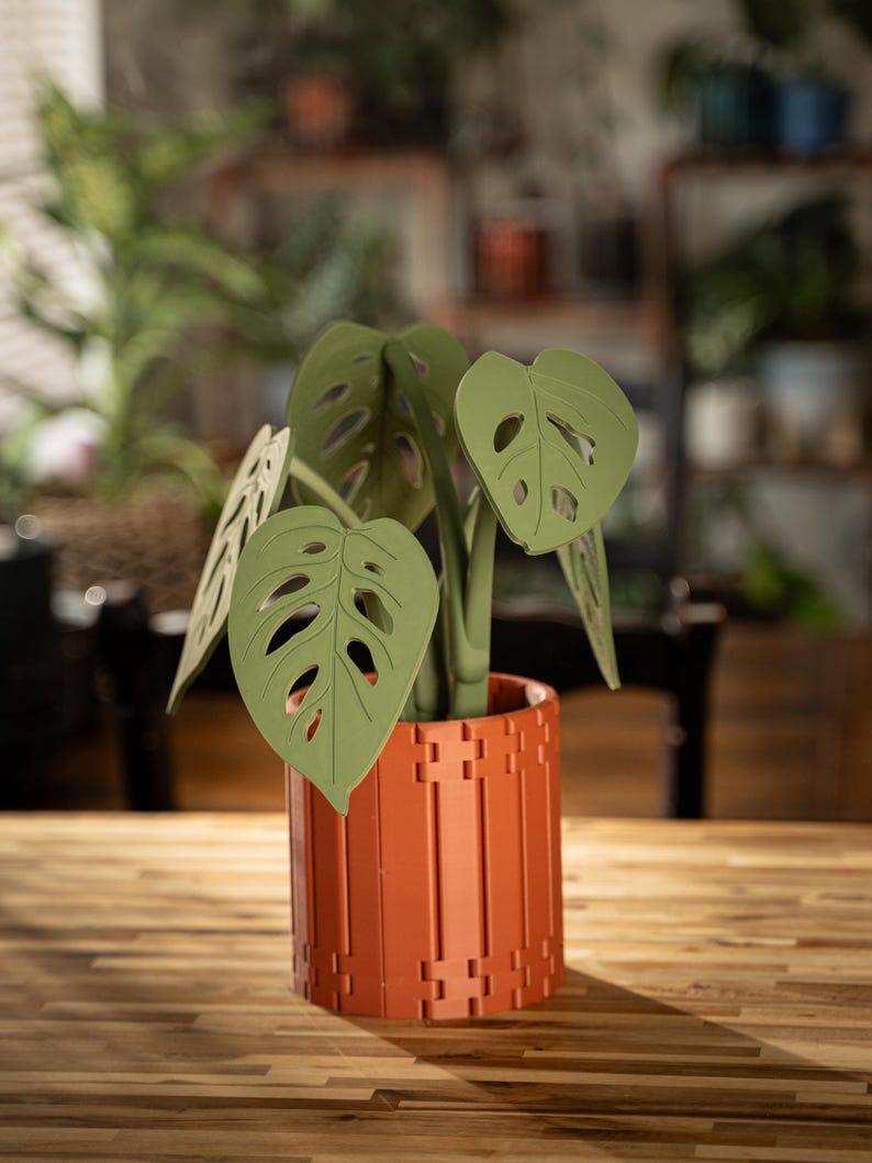 May include: A decorative artificial plant with green leaves and a terracotta-colored pot. The plant features large, perforated leaves and is set in a cylindrical pot with a geometric design. The pot is made of a reddish-brown material and sits on a wooden surface.