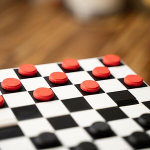May include: A close-up of a checkerboard with red and black checkers. The board is white with black squares. The red checkers are on the top half of the board, while the black checkers are on the bottom half.