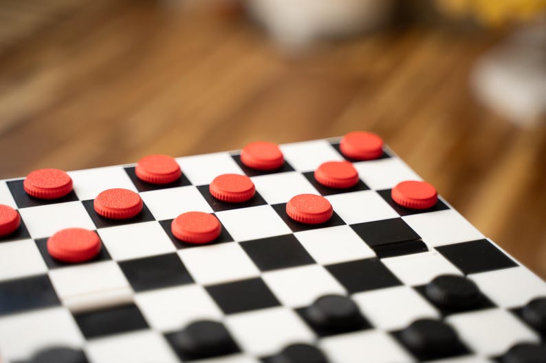 May include: A close-up of a checkerboard with red and black checkers. The board is white with black squares. The red checkers are on the top half of the board, while the black checkers are on the bottom half.