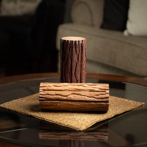 May include: Two decorative wooden log-shaped items. One is a tall, cylindrical piece with a dark brown, textured bark-like surface. The other is a shorter, rectangular piece with a lighter brown, detailed wood grain. Both are on a gold mat.
