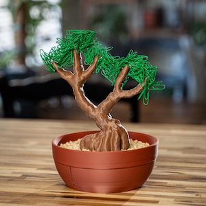 Könnte beinhalten: Eine braune Bonsai-Baumskulptur mit grünen Büroklammer-Blättern steht in einem terrakottafarbenen Topf. Der Baumstamm hat ein strukturiertes Aussehen, und der Topf ist mit kleinen, hellfarbigen Kieselsteinen gefüllt. Der Bonsai steht auf einer Holzoberfläche.