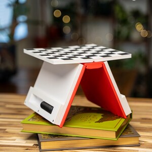 May include: A white and red folding chess board stand with a black and white checkered surface. The stand is placed on top of two books with green and gold covers. The stand has a black, cylindrical component on the side.