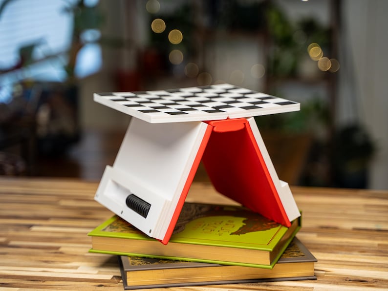May include: A white and red folding chess board stand with a black and white checkered surface. The stand is placed on top of two books with green and gold covers. The stand has a black, cylindrical component on the side.