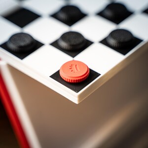 May include: Close-up of a checkerboard with black and white squares and black checkers. A red checker is on the corner square. The checkerboard is part of a red and white game set.