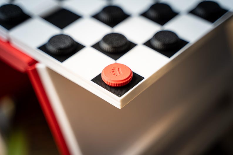 May include: Close-up of a checkerboard with black and white squares and black checkers. A red checker is on the corner square. The checkerboard is part of a red and white game set.