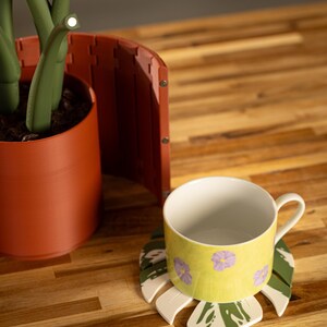 May include: A yellow ceramic mug with purple floral designs sits on a leaf-shaped coaster with green and white accents. The mug is on a wooden surface, near a terracotta-colored plant pot and a decorative screen.