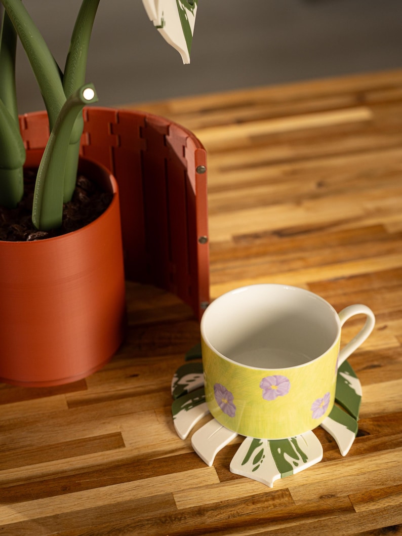 May include: A yellow ceramic mug with purple floral designs sits on a leaf-shaped coaster with green and white accents. The mug is on a wooden surface, near a terracotta-colored plant pot and a decorative screen.