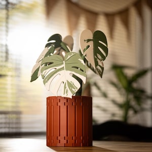 Op de afbeelding: Een decoratieve houten plant met grote, gespleten bladeren in groen en wit, in een terracotta pot met een geometrisch patroon. De plant is een gestileerde Monstera, ideaal voor interieurdecoratie.