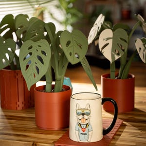May include: A white ceramic mug with a black handle and a bulldog graphic wearing sunglasses and a shirt that says "MUSIC". The mug sits on a red coaster next to decorative paper plants in terracotta pots.