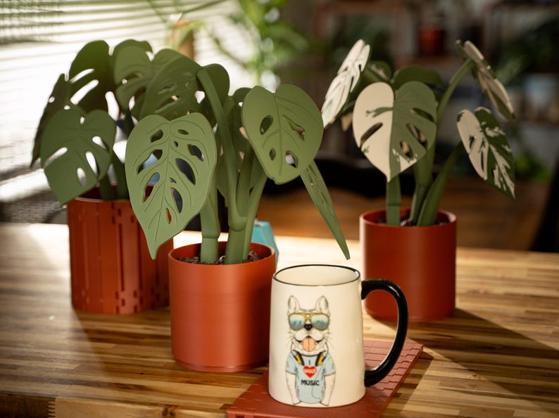 May include: A white ceramic mug with a black handle and a bulldog graphic wearing sunglasses and a shirt that says "MUSIC". The mug sits on a red coaster next to decorative paper plants in terracotta pots.