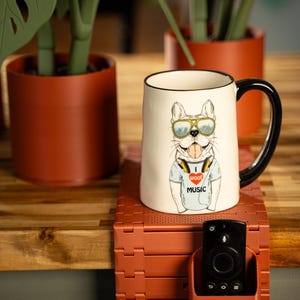 May include: A ceramic mug with a black handle and rim features a cartoon bulldog wearing sunglasses and headphones. The bulldog is wearing a t-shirt with the text "I woof music". The mug is on a red-brown stand with a remote control holder.