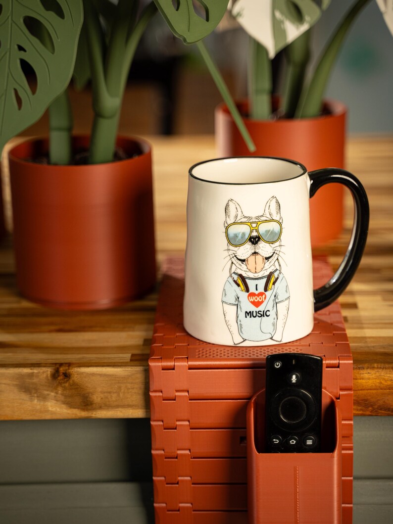 May include: A ceramic mug with a black handle and rim features a cartoon bulldog wearing sunglasses and headphones. The bulldog is wearing a t-shirt with the text "I woof music". The mug is on a red-brown stand with a remote control holder.