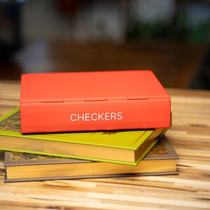 May include: A red checkerboard game box with the word "CHECKERS" in white lettering is stacked on top of two vintage books with green and gold covers. The books are stacked on a wooden surface.