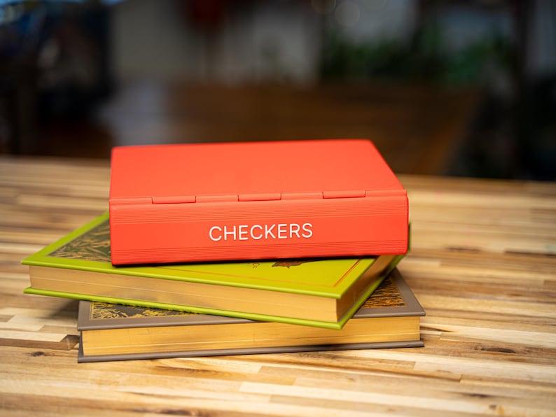 May include: A red checkerboard game box with the word "CHECKERS" in white lettering is stacked on top of two vintage books with green and gold covers. The books are stacked on a wooden surface.