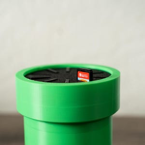 以下が含まれることがあります： 鮮やかな緑色の円筒形の容器で、ビデオゲームのパイプに似ており、内側は黒色です。Nintendo Switchのゲームカートリッジが中に入っています。容器はプラスチック製で、滑らかな仕上がりです。