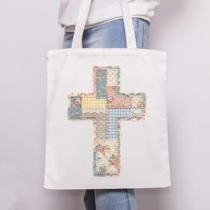 K&ouml;nnte beinhalten: Wei&szlig;e Tragetasche mit einem Patchwork-Kreuz-Design. Das Kreuz besteht aus verschiedenen floralen und gemusterten Stoffen in Pastellt&ouml;nen, darunter Rosa, Blau, Gelb und Gr&uuml;n. Die Tasche hat zwei wei&szlig;e Griffe.