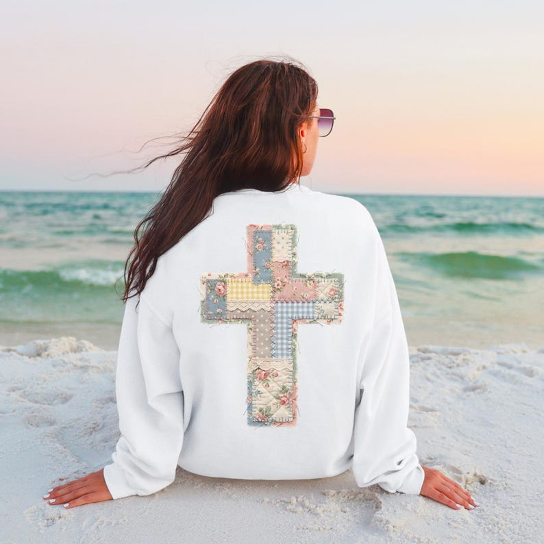 K&ouml;nnte beinhalten: Wei&szlig;es Sweatshirt mit einem Patchwork-Kreuz-Design auf dem R&uuml;cken. Das Kreuz besteht aus verschiedenen Stoffmustern, darunter Blumenmuster, Vichy-Muster und unifarbene Stoffe. Das Sweatshirt wird an einem Strand mit dem Meer im Hintergrund getragen.