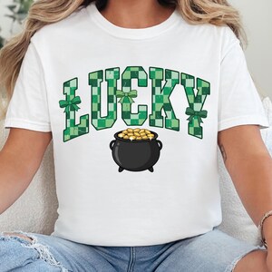 May include: White t-shirt with the word "LUCKY" in green checkered letters, green bows, and a black pot filled with gold coins. A festive design for St. Patrick's Day.