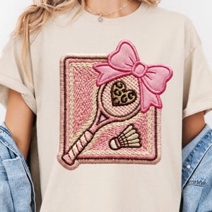 May include: A beige t-shirt featuring a large embroidered patch of a tennis racket with a leopard print heart in the center, a pink bow at the top, and a shuttlecock at the bottom, all set against a square pink and cream textured background.