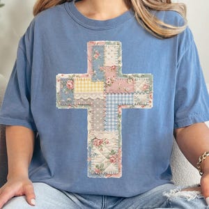 K&ouml;nnte beinhalten: Hellblaues T-Shirt mit einem Patchwork-Kreuz-Design. Das Kreuz besteht aus verschiedenen Stoffmustern, darunter Blumenmuster, Vichy-Karo und Spitze, in Pastellfarben. Das Design befindet sich mittig auf der Vorderseite des Shirts.