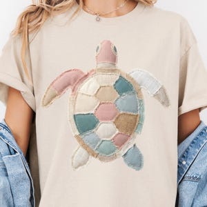 May include: Beige t-shirt featuring a patchwork turtle design. The turtle has a pink head, light blue, pink, and beige shell, and light blue and beige flippers. The design is made of various fabric patches.