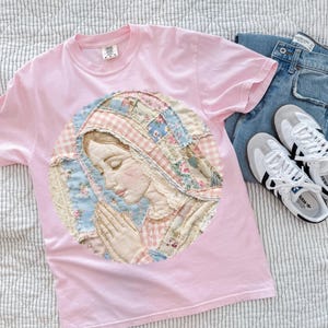 Könnte beinhalten: Hellrosa T-Shirt mit einem kreisförmigen Applikation, die eine betende Figur darstellt. Die Applikation besteht aus Patchwork-Stoff in Pastellfarben. Blaue Jeans und weiße Turnschuhe mit schwarzen Streifen sind ebenfalls zu sehen.