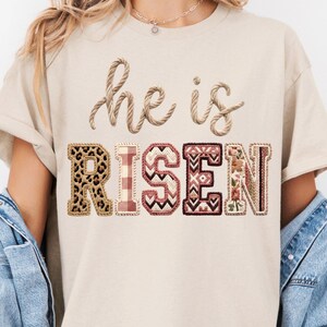 May include: Beige t-shirt with the words "he is RISEN" in a decorative design. The word "he is" is in a rope-like font, and "RISEN" features a leopard print, plaid, geometric, and floral pattern.