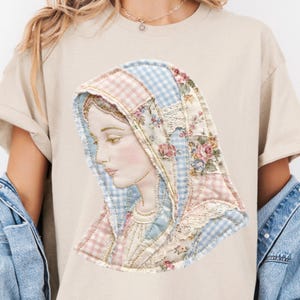 May include: Beige t-shirt featuring a detailed portrait of a woman with a head covering. The portrait is made of fabric patches in pink, blue, and floral patterns. The woman's face is delicately drawn with soft colors.