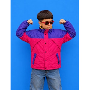 May include: A color-blocked jacket featuring a purple upper section and a vibrant pink lower section, paired with red-framed sunglasses. The jacket includes a high collar with a yellow inner lining, long sleeves, and a central button-down closure.