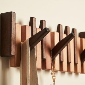 May include: A wooden wall-mounted coat rack with a modern design. The rack features alternating dark brown and light wood blocks, with angled pegs extending outward. A beige and brown striped strap and a gold necklace hang from the pegs.
