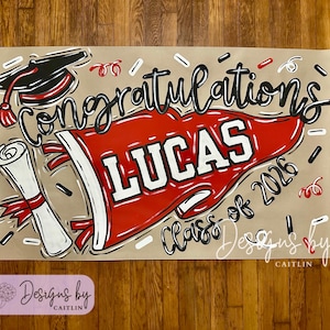 May include: A graduation banner with the word "Congratulations" in black script. A red pennant with the name "LUCAS" in white lettering is featured. The banner also includes a graduation cap, diploma, and the text "Class of 2026".