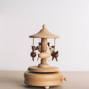 May include: A wooden carousel music box featuring a light-toned circular base with a winding knob on the side. The central pillar supports a conical roof and four small, alternating light and dark brown wooden horses suspended by thin metal rods.