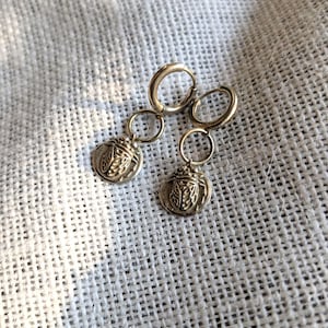 May include: A pair of gold-colored hoop earrings. Each earring features a small, circular charm with a detailed insect design. The earrings are displayed on a textured, off-white fabric.