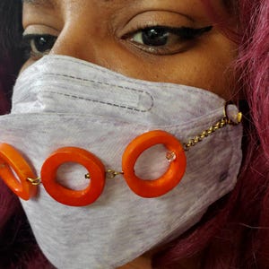 Puede incluir: Una mascarilla facial gris claro con detalles circulares naranjas conectados por una cadena dorada. La mascarilla tiene un diseño decorativo y se lleva en la cara. La persona tiene la piel oscura y el pelo rojo.