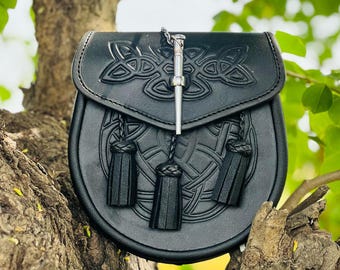 May include: A black leather sporran with a Celtic knot design and three black tassels. A silver chain with a decorative silver pin is attached to the sporran.