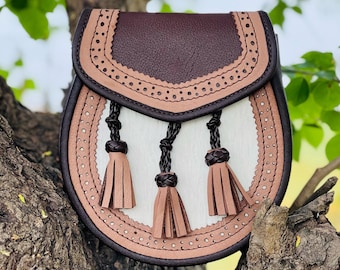 Puede incluir: Un bolso de cuero marrón con una solapa de cuero blanco y tres borlas marrones. El bolso tiene un borde decorativo de pequeños agujeros y costuras.