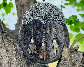 May include: A traditional Scottish sporran with a silver cantle featuring Celtic knotwork and a stag design. The fur pouch is grey and brown, with three decorative tassels hanging from chains. The sporran is displayed outdoors, resting on a tree branch.