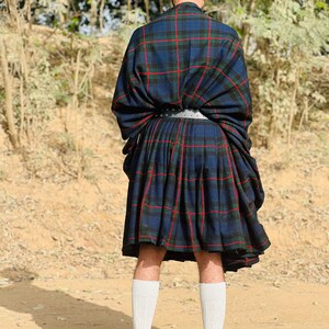 May include: A person wearing a blue and red plaid kilt with a black leather belt and black boots. The kilt is folded over at the waist and the person is standing on a dirt path.
