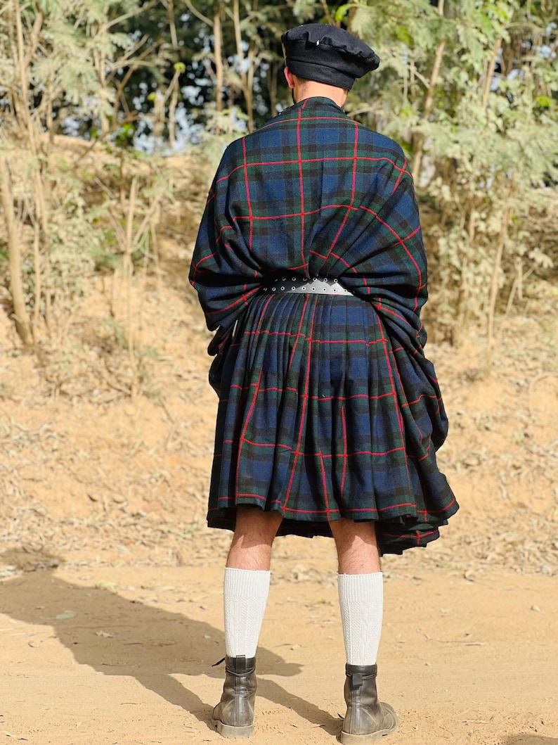 May include: A person wearing a blue and red plaid kilt with a black leather belt and black boots. The kilt is folded over at the waist and the person is standing on a dirt path.
