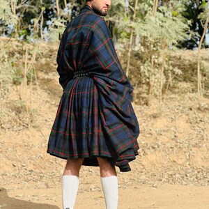 May include: A man wearing a black beret, a black belt with silver studs, and a blue, green, and red plaid kilt. He is standing on a dirt path with trees in the background.