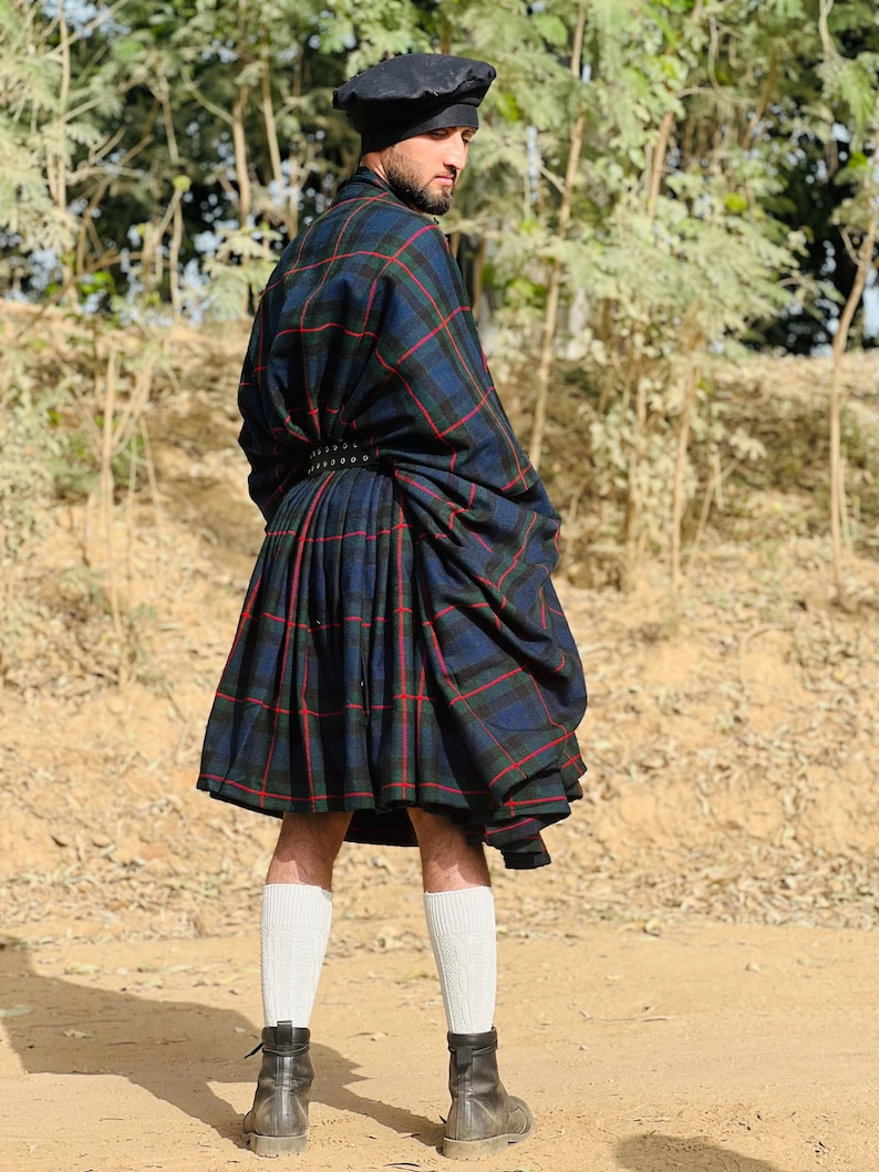 May include: A man wearing a black beret, a black belt with silver studs, and a blue, green, and red plaid kilt. He is standing on a dirt path with trees in the background.