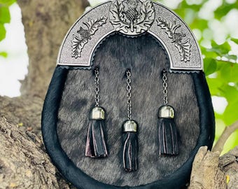 May include: A traditional Scottish sporran with a silver cantle featuring thistle designs. The sporran has a gray fur body, black leather trim, and three black tassels with silver caps hanging from chains.